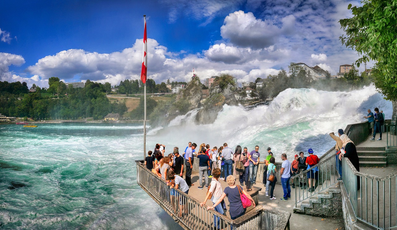 In this image, a group of individuals is depicted standing on a staircase. Adjacent to them, a fence, a flag, a body of water, trees, buildings, and a cloudy sky are visible.\n
