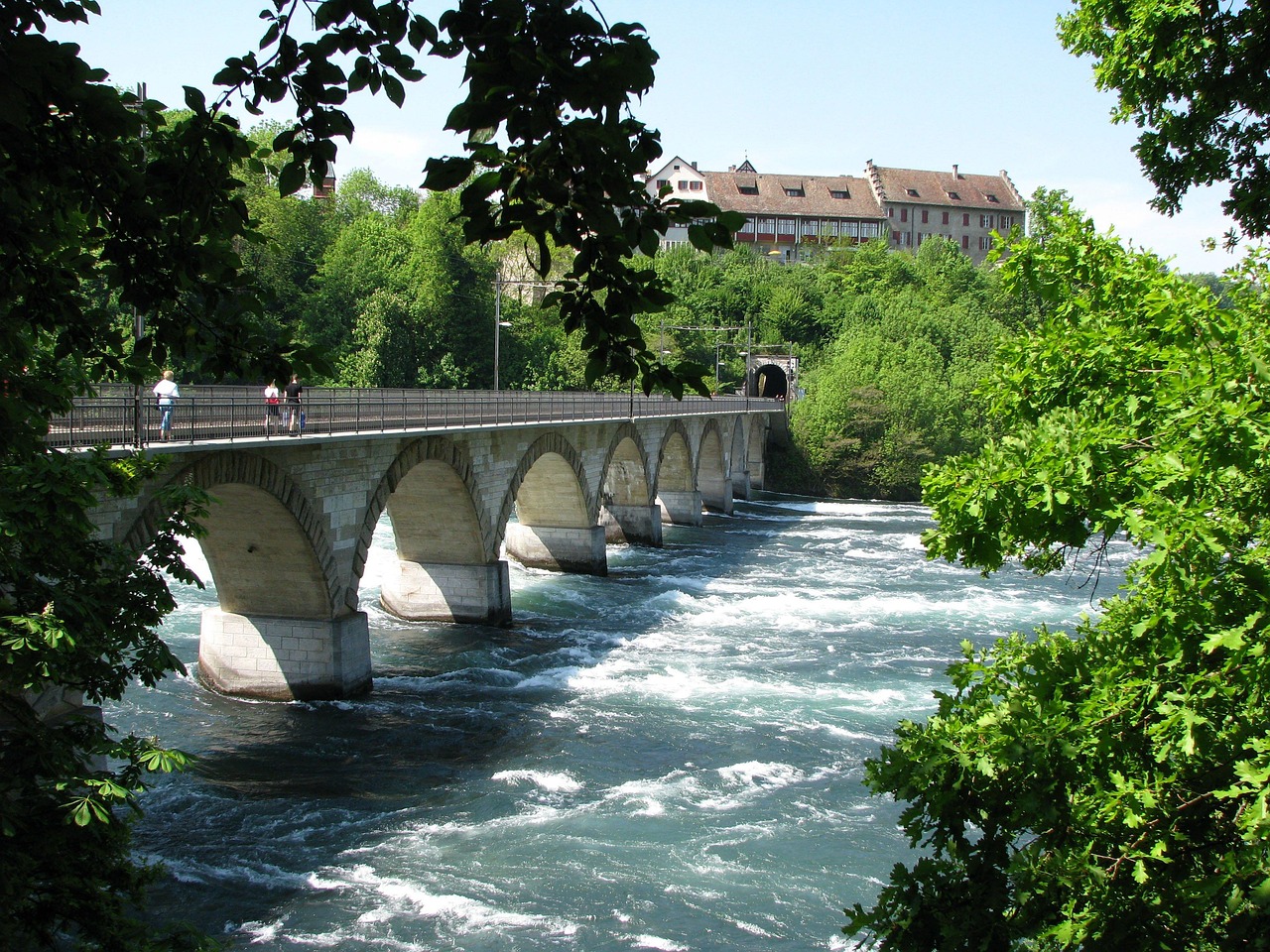 In this image, a bridge is visible, with a few individuals traversing its surface. The water surrounding the bridge is prominent, and a pole stands tall. The background of the image encompasses a verdant landscape, featuring trees, buildings, and a vast expanse of sky.\n
